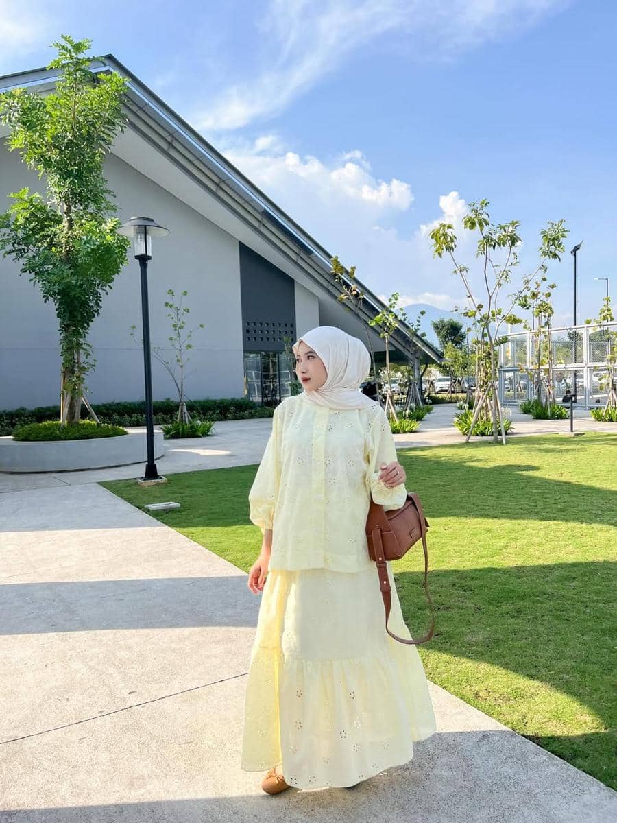 Seorang wanita berhijab mengenakan outfit kuning pastel dengan tas cokelat berjalan di area taman modern di bawah langit cerah.