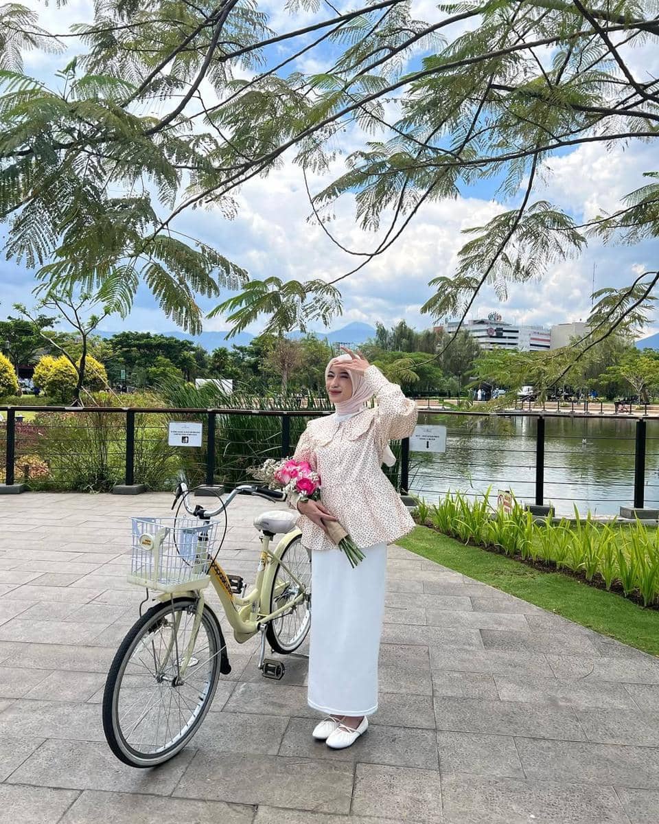 Seorang wanita berhijab bergaya santai dengan sepeda dan buket bunga di tepi danau, dikelilingi pepohonan dan langit cerah.