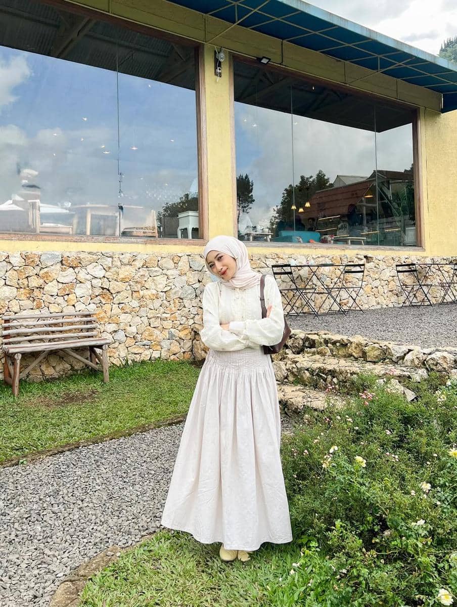 Seorang wanita berhijab mengenakan blus putih dan rok panjang berwarna krem berpose di depan kafe dengan dinding batu dan taman hijau.
