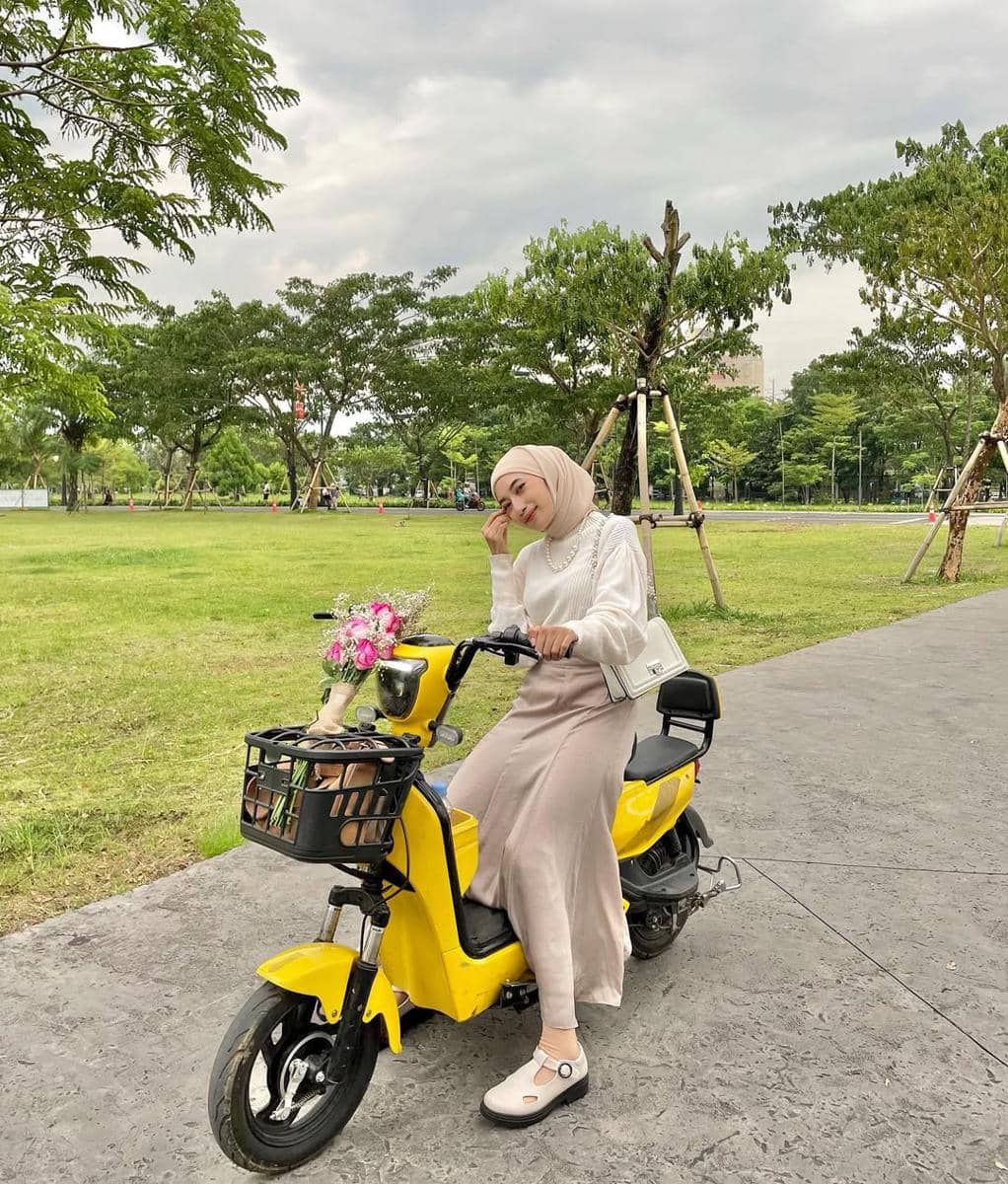 Seorang wanita berhijab duduk di atas motor listrik kuning dengan keranjang berisi bunga di taman hijau pada hari berawan.
