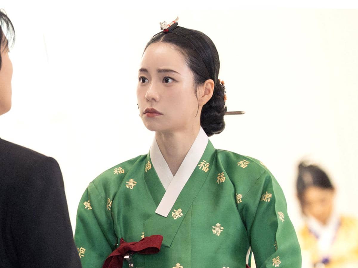 Seorang wanita mengenakan hanbok hijau tradisional dengan rambut disanggul rapi sedang berbicara dengan seseorang di depannya.
