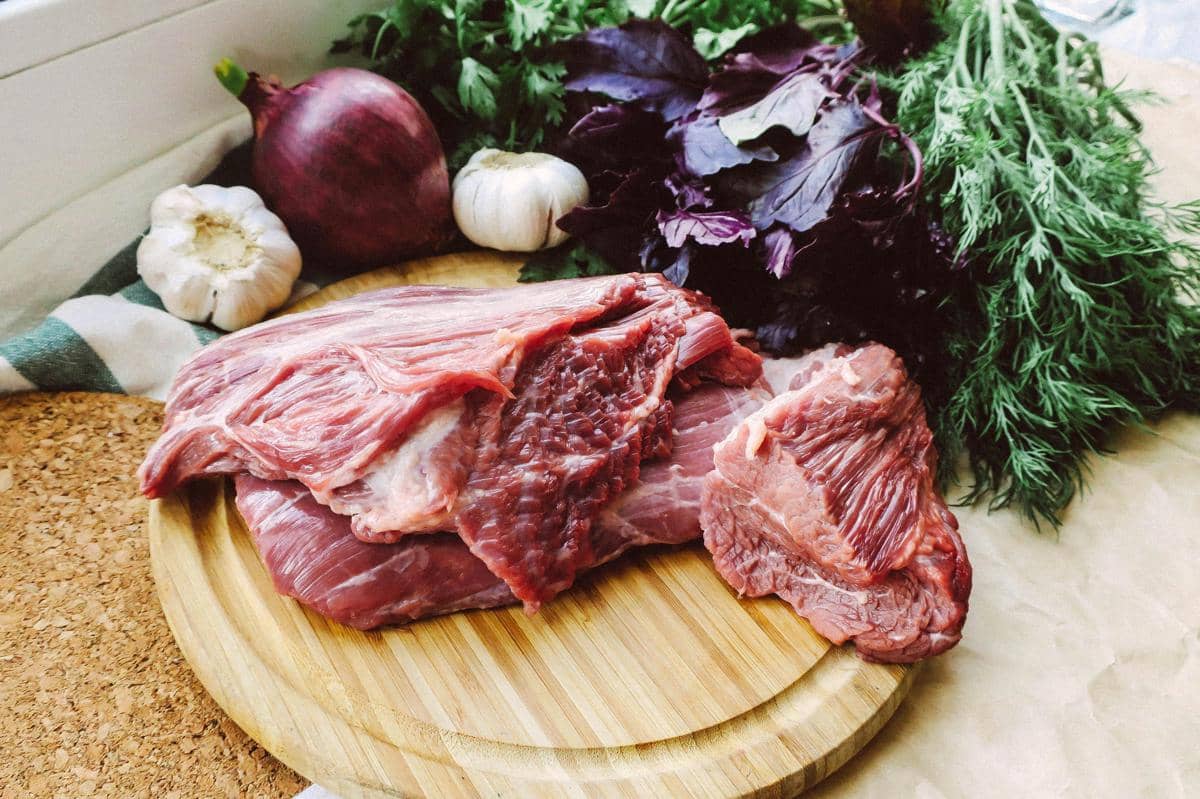 Potongan daging kambing segar di atas talenan kayu dengan bawang putih, bawang merah, dan daun rempah di sekelilingnya.