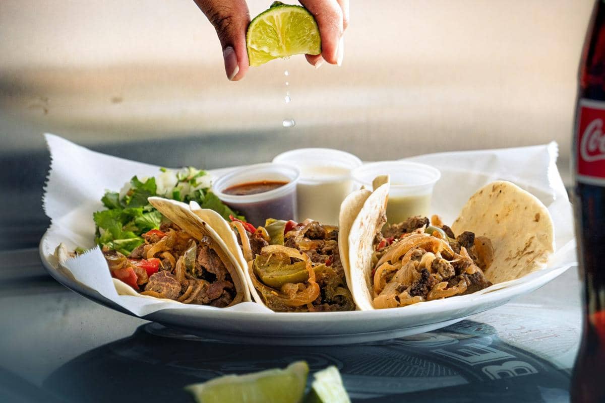 Tiga taco fajita steak dengan irisan daging sapi, paprika, dan bawang disajikan di piring, diberi perasan jeruk nipis segar di atasnya.