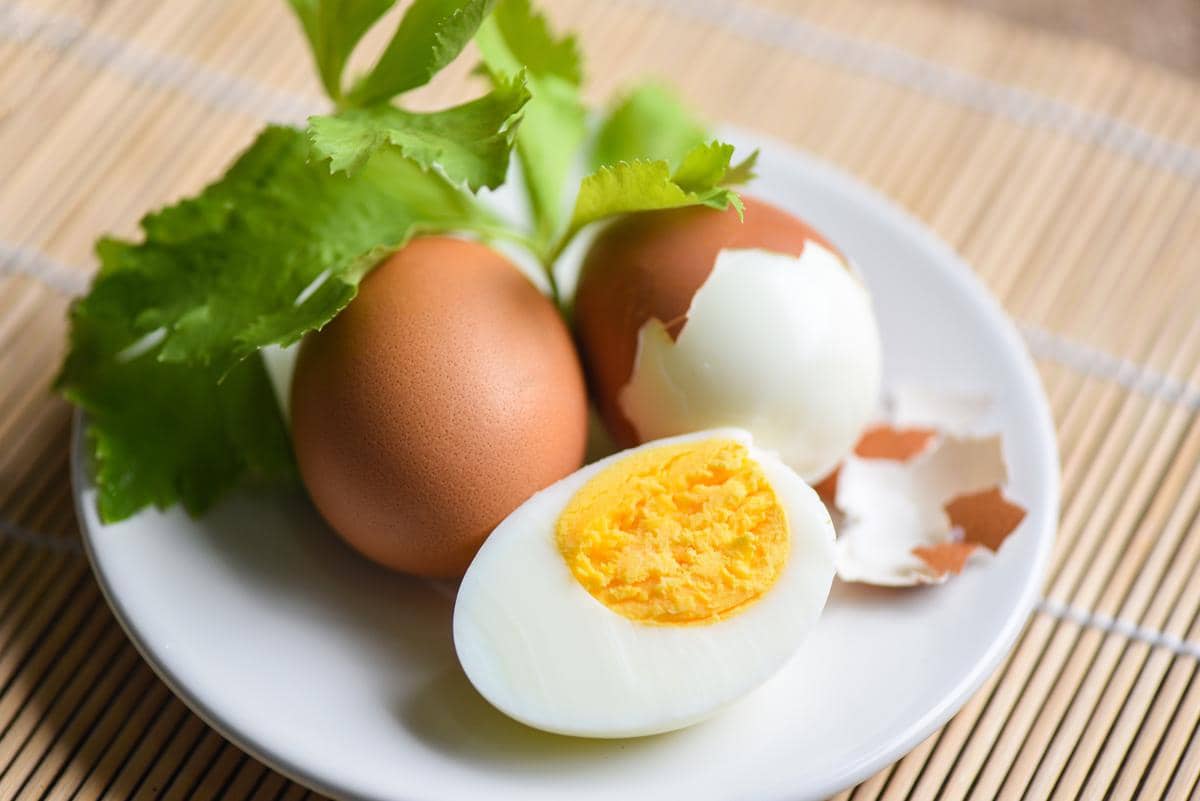 Telur rebus setengah dikupas dan telur utuh berwarna cokelat disajikan di atas piring putih dengan hiasan daun seledri segar.