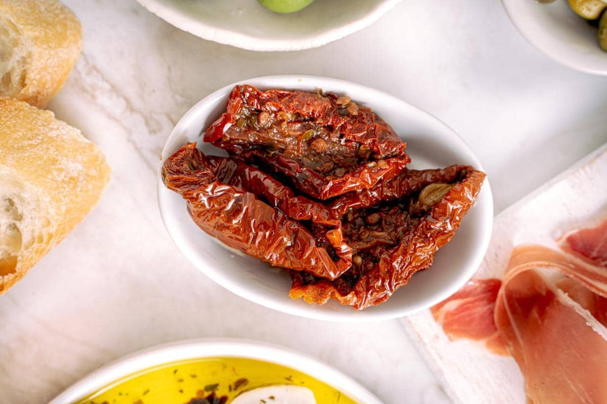 Beberapa potong tomat kering berwarna merah tua disajikan dalam mangkuk putih di atas meja marmer bersama roti dan daging asap.