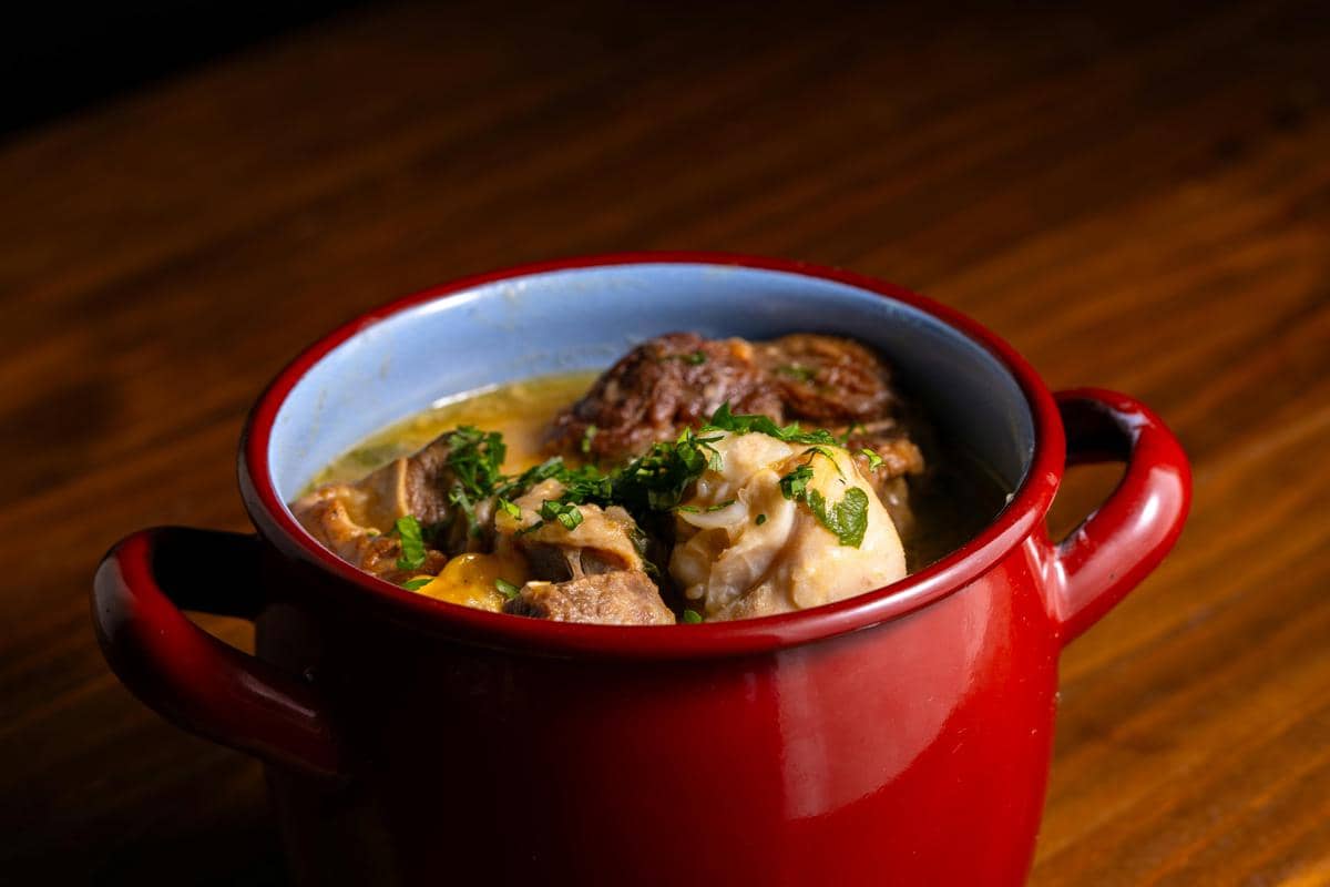 Semangkuk asam-asam daging disajikan dalam mangkuk merah dengan kuah berwarna kekuningan dan taburan daun hijau segar di atasnya.