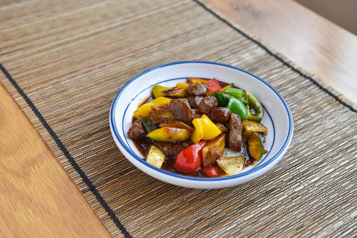 Semangkuk daging sapi lada hitam dengan potongan paprika merah, kuning, dan hijau disajikan di atas meja beralaskan anyaman bambu.