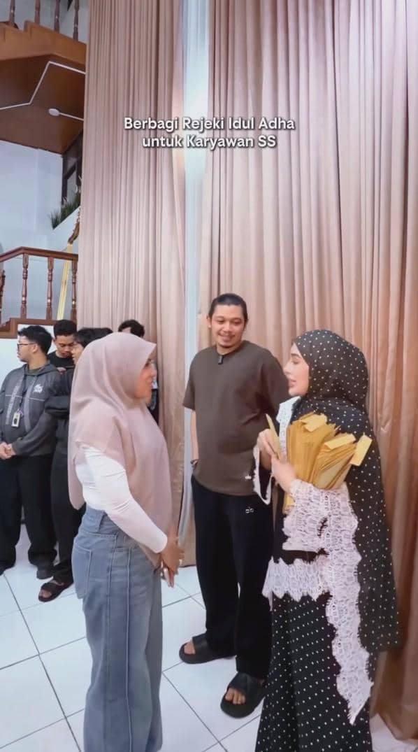 Seorang wanita berhijab hitam berbaju polkadot membagikan amplop THR Iduladha kepada karyawan yang berbaris di dalam ruangan.