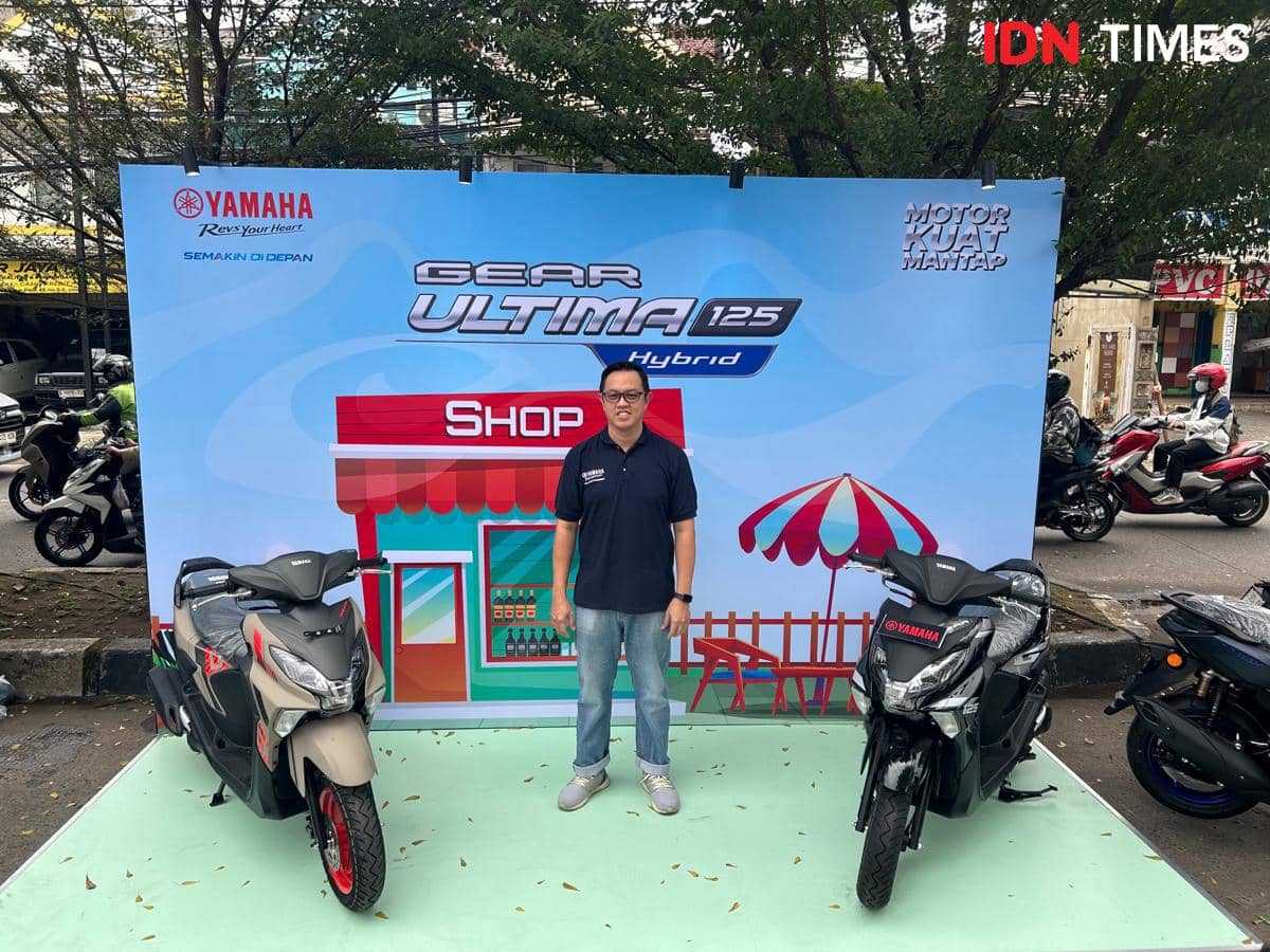 Seorang pria berdiri di antara dua motor Yamaha Gear Ultima 125 Hybrid dalam acara karnaval di Depok dengan latar promosi produk.