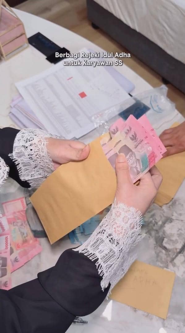Seseorang mengenakan pakaian berlengan renda putih memasukkan uang rupiah ke dalam amplop cokelat untuk dibagikan sebagai THR Iduladha.