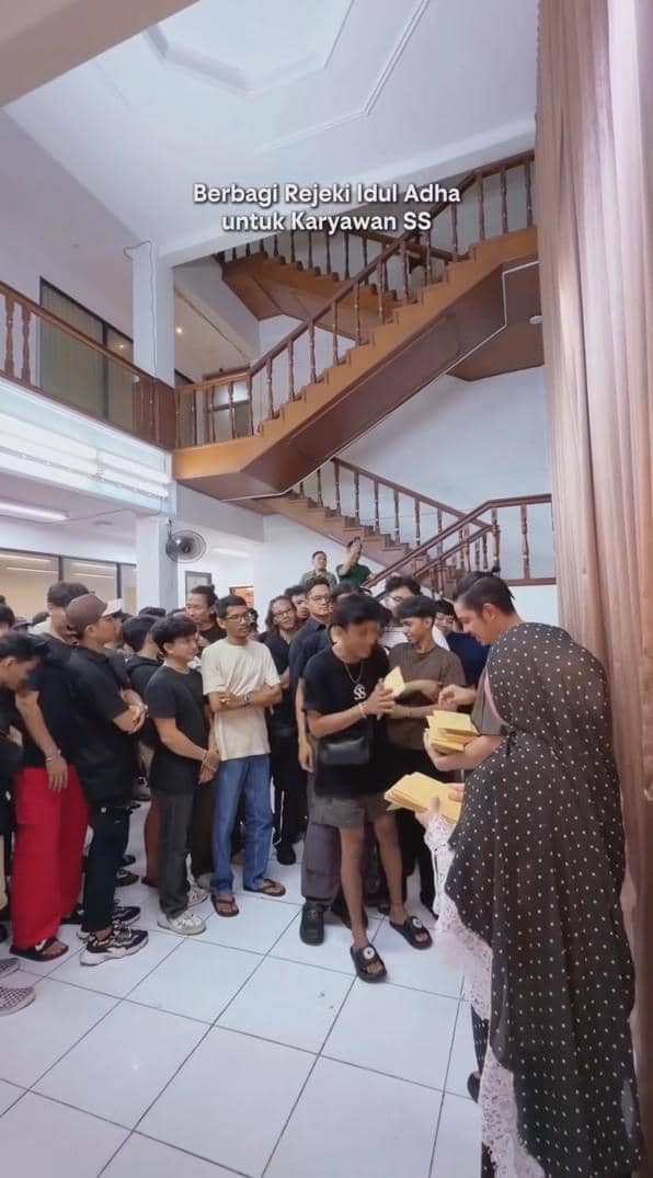 Seorang wanita berhijab membagikan amplop berisi THR Iduladha kepada sejumlah karyawan yang berbaris di dalam gedung bertingkat.
