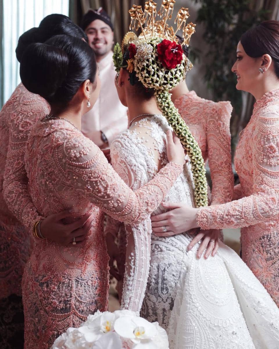 Pengantin wanita mengenakan kebaya putih berhias sanggul dan bunga melati panjang, dikelilingi para bridesmaid berkebaya merah muda.