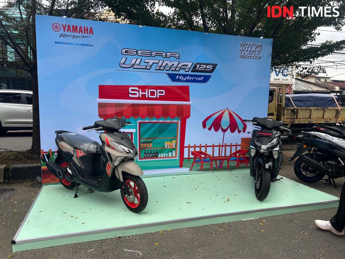 Dua motor Yamaha Gear Ultima 125 Hybrid dipamerkan di area luar ruangan dengan latar belakang bertema toko dan kafe kecil.