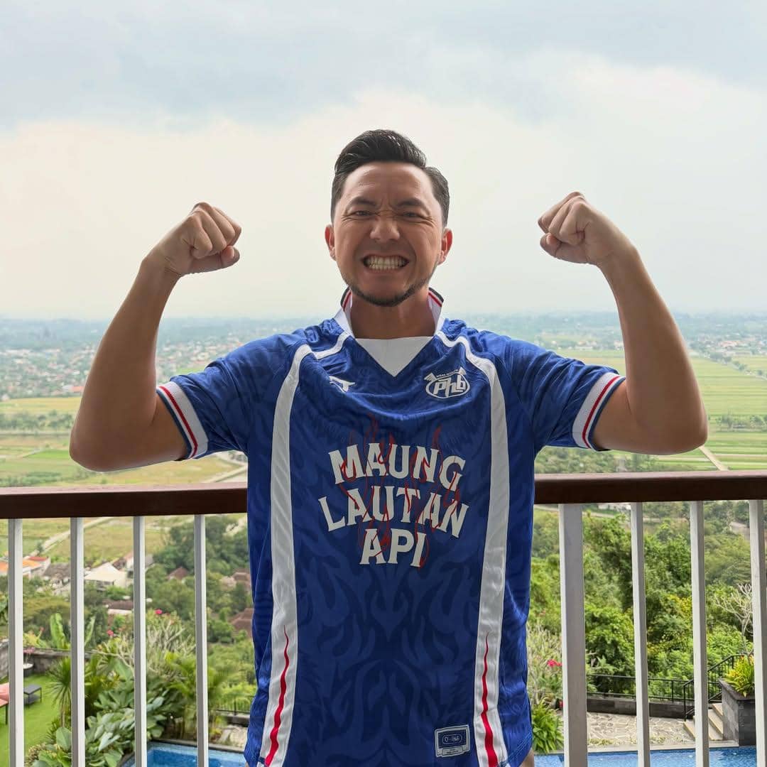 Seorang pria mengenakan jersey Persib Bandung berwarna biru dengan tulisan Maung Lautan Api sambil mengangkat kedua tangannya dengan ekspresi gembira.