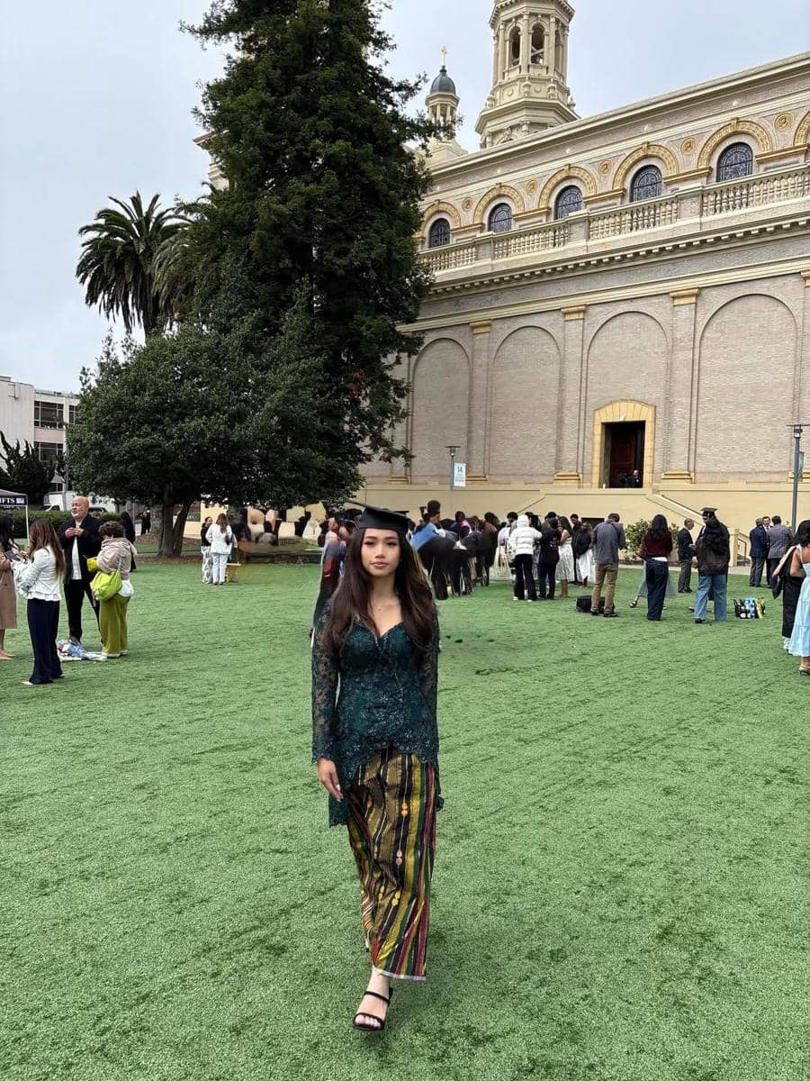 Seorang perempuan mengenakan kebaya hijau dan kain batik berjalan di area rumput dengan latar gedung bergaya klasik saat acara wisuda.