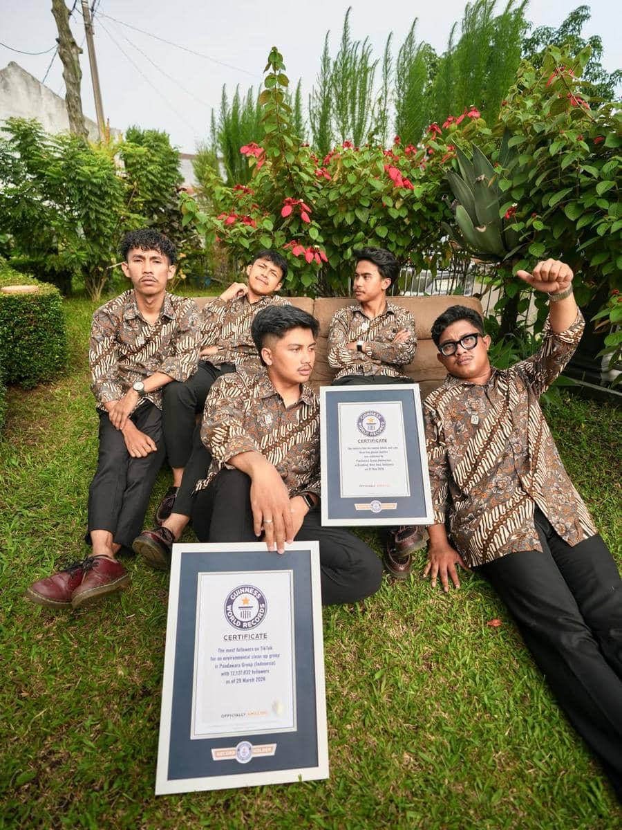 Lima pria mengenakan batik duduk di taman sambil memegang dua sertifikat Guinness World Records dengan latar pepohonan hijau.