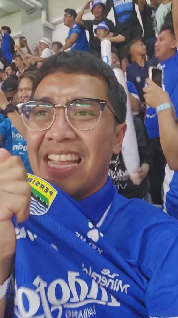 Seorang pria mengenakan jersey biru Persib Bandung bersorak gembira di tengah kerumunan suporter di stadion.