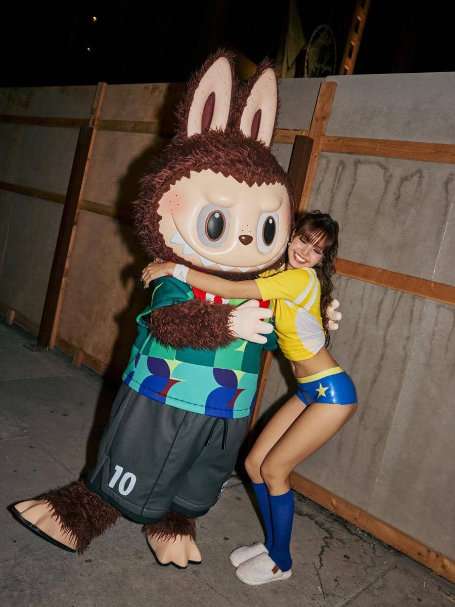 Seorang perempuan mengenakan kostum bertema sepak bola berwarna kuning dan biru memeluk karakter maskot besar berbentuk kelinci di area belakang panggung.