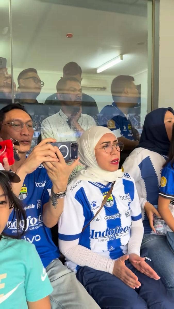 Sejumlah orang mengenakan jersey Persib Bandung duduk di tribun menonton pertandingan sambil bersorak dan merekam momen dengan ponsel.