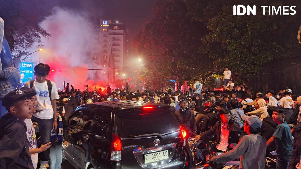 Kerumunan warga dan kendaraan memenuhi area Tugu Kujang Bogor pada malam hari dengan asap merah di latar belakang.
