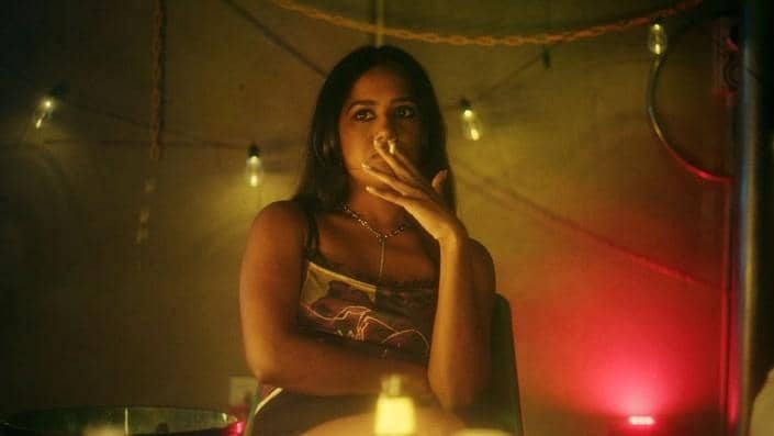 Seorang perempuan duduk di ruangan bercahaya kuning dan merah sambil merokok, dengan ekspresi tenang dan suasana misterius.