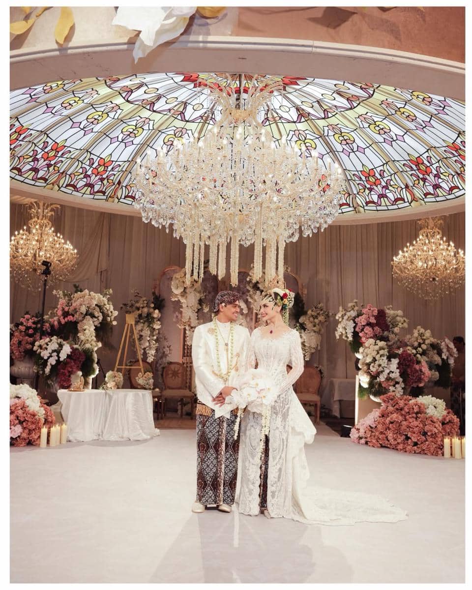Pasangan pengantin mengenakan busana adat Jawa berdiri di pelaminan mewah dengan hiasan bunga dan lampu gantung kristal besar.