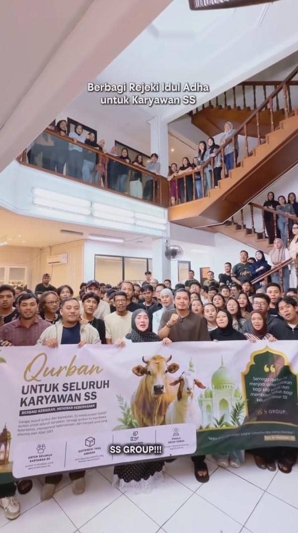 Sekelompok besar karyawan berkumpul di dalam gedung bertingkat memegang spanduk bertema Iduladha dengan tulisan Qurban untuk seluruh karyawan SS.