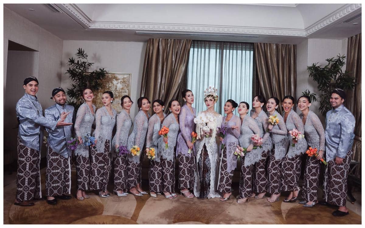 Pengantin wanita mengenakan kebaya putih dan siger berdiri bersama para bridesmaid dan groomsman dalam busana tradisional berwarna senada.