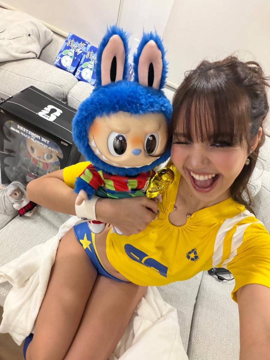 Seorang perempuan mengenakan kostum kuning dan biru berpose ceria sambil memeluk boneka berkepala kelinci biru di ruang santai.