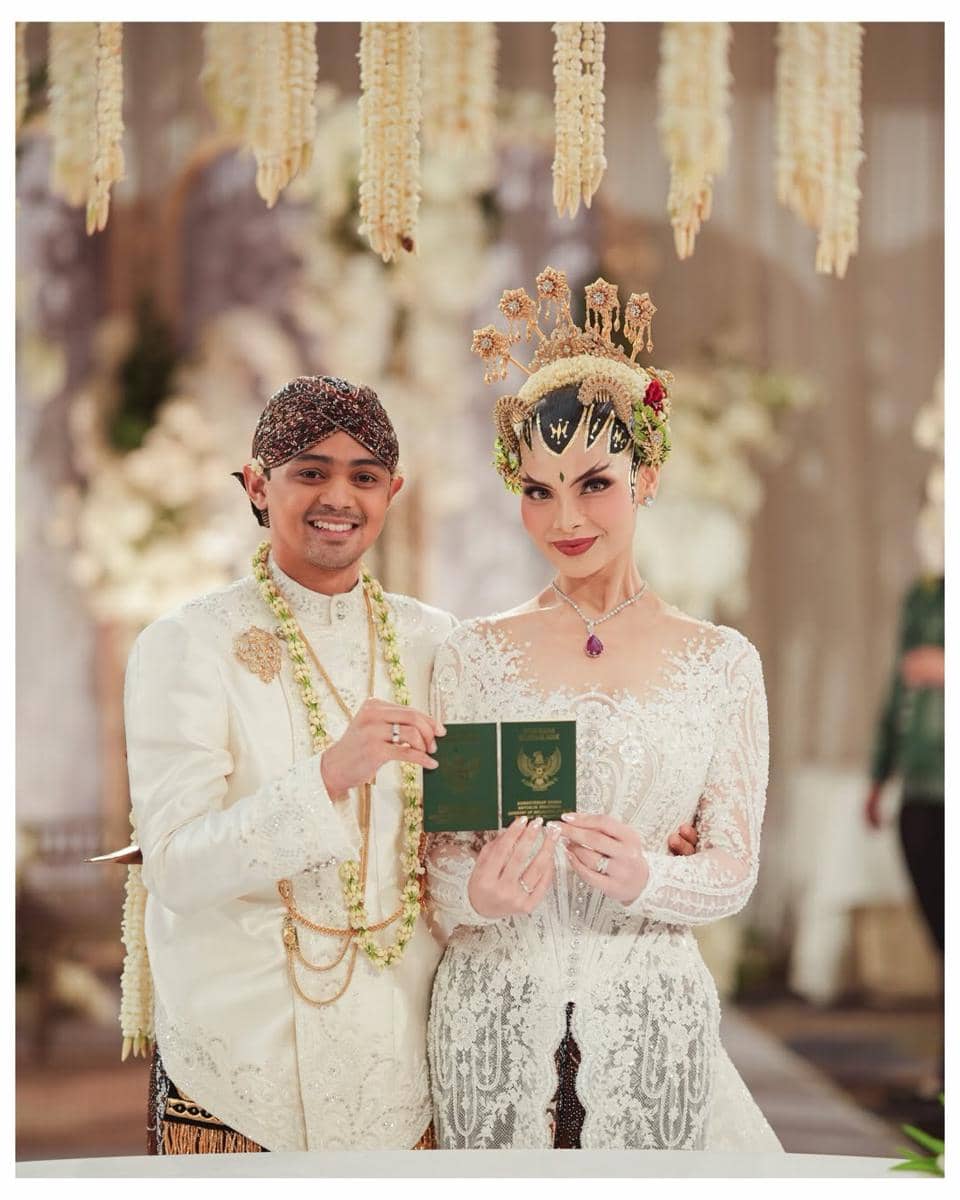 Pasangan pengantin mengenakan busana adat Jawa berwarna putih sambil memegang buku nikah di acara pernikahan yang elegan.