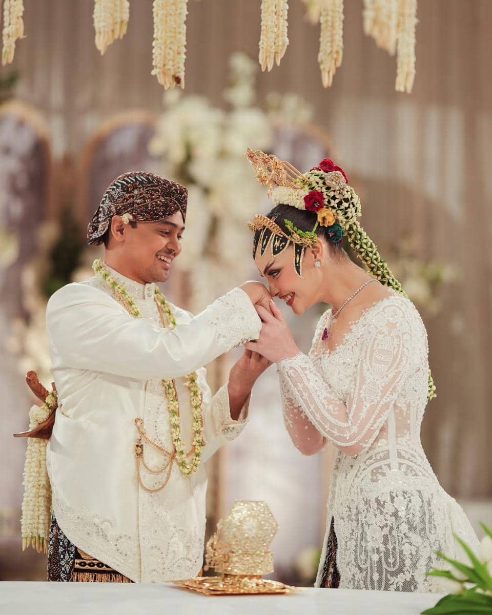 Pasangan pengantin mengenakan busana adat Jawa berwarna putih dalam prosesi pernikahan dengan dekorasi bunga melati di latar belakang.