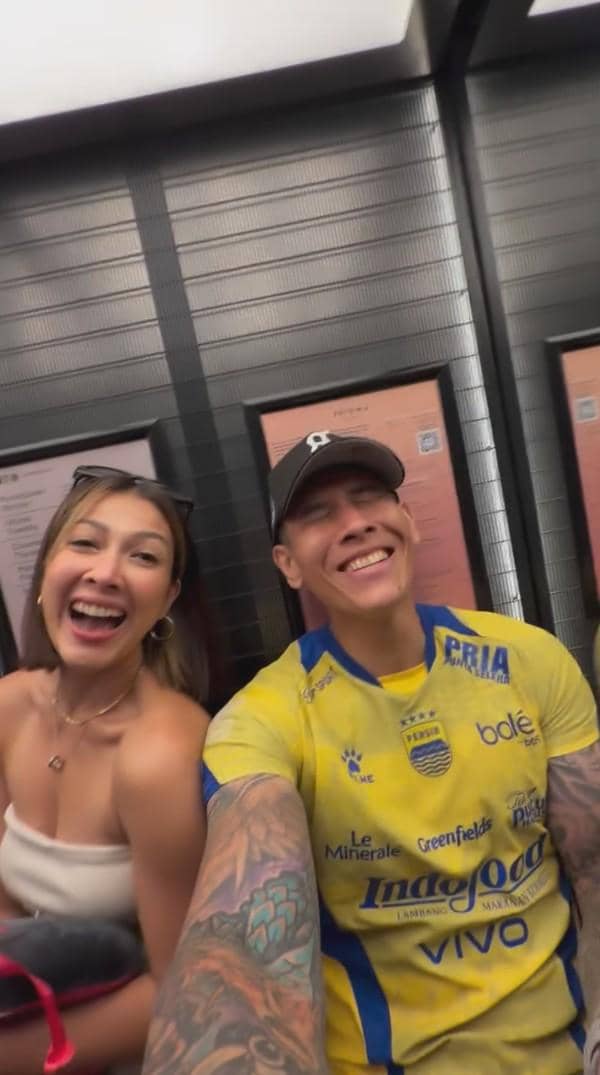 Seorang pria mengenakan jersey Persib Bandung berwarna kuning dan seorang wanita tersenyum lebar saat berfoto bersama di dalam lift.