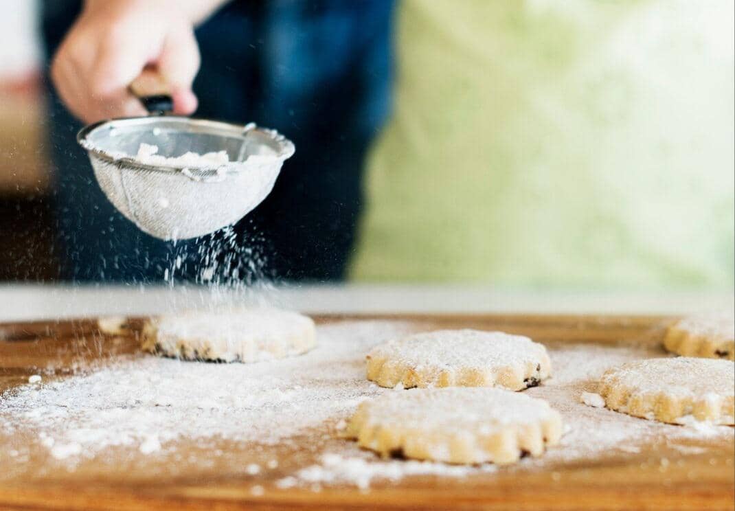 Tangan menaburkan gula halus menggunakan saringan di atas beberapa kue kering berbentuk bulat di atas meja kayu.