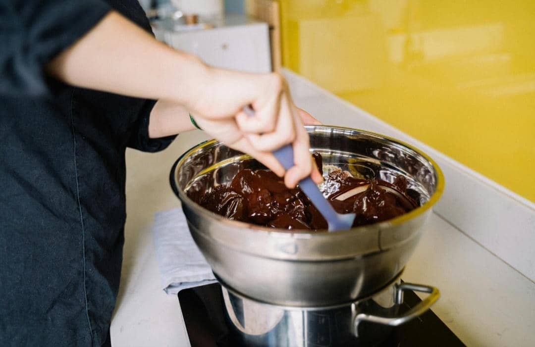 Seseorang mengaduk cokelat leleh di dalam mangkuk logam menggunakan metode double boiler di dapur modern.