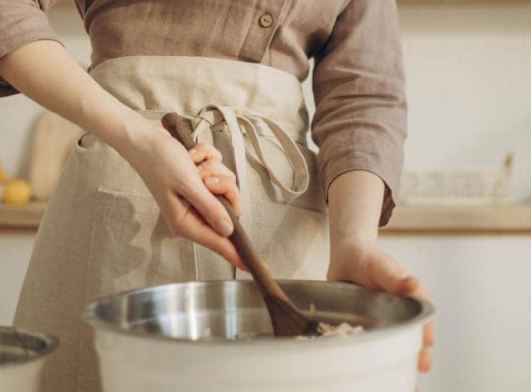 Seseorang mengenakan celemek berwarna krem sedang mengaduk adonan di dalam mangkuk logam menggunakan spatula kayu di dapur.