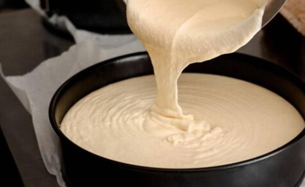 Adonan cheesecake lembut dituangkan ke dalam loyang bulat hitam yang sudah dilapisi kertas roti sebelum dipanggang.