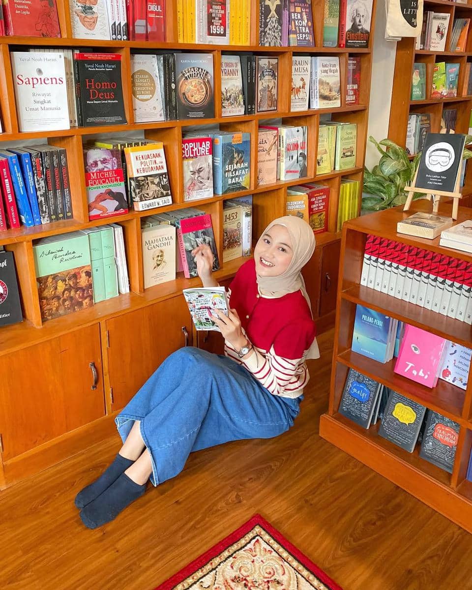 Seorang wanita berhijab mengenakan sweater merah dan rok denim duduk di lantai perpustakaan sambil membaca buku di depan rak penuh buku.