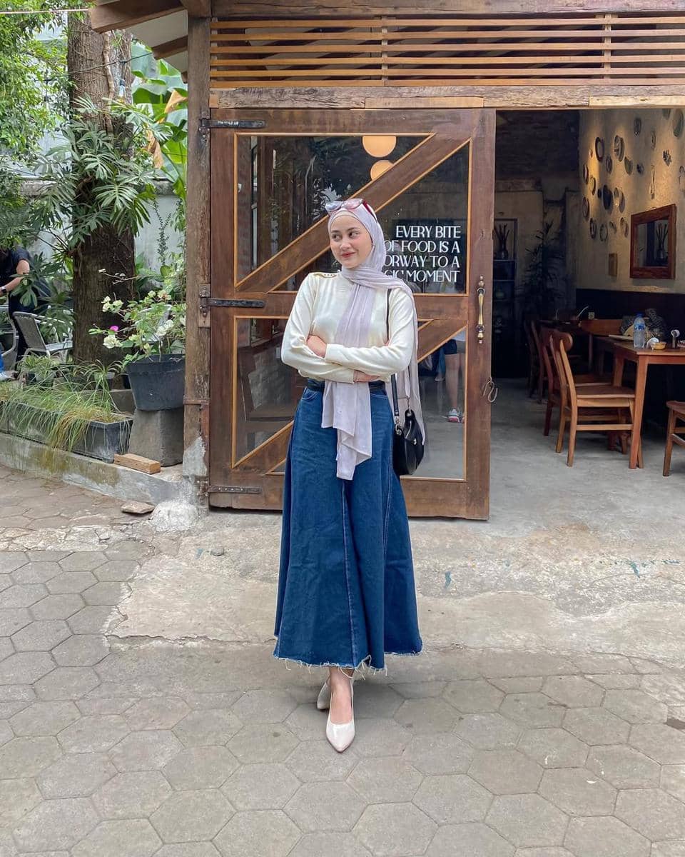 Seorang wanita berhijab mengenakan atasan putih dan rok denim panjang berdiri di depan kafe bergaya rustic dengan pintu kayu kaca.