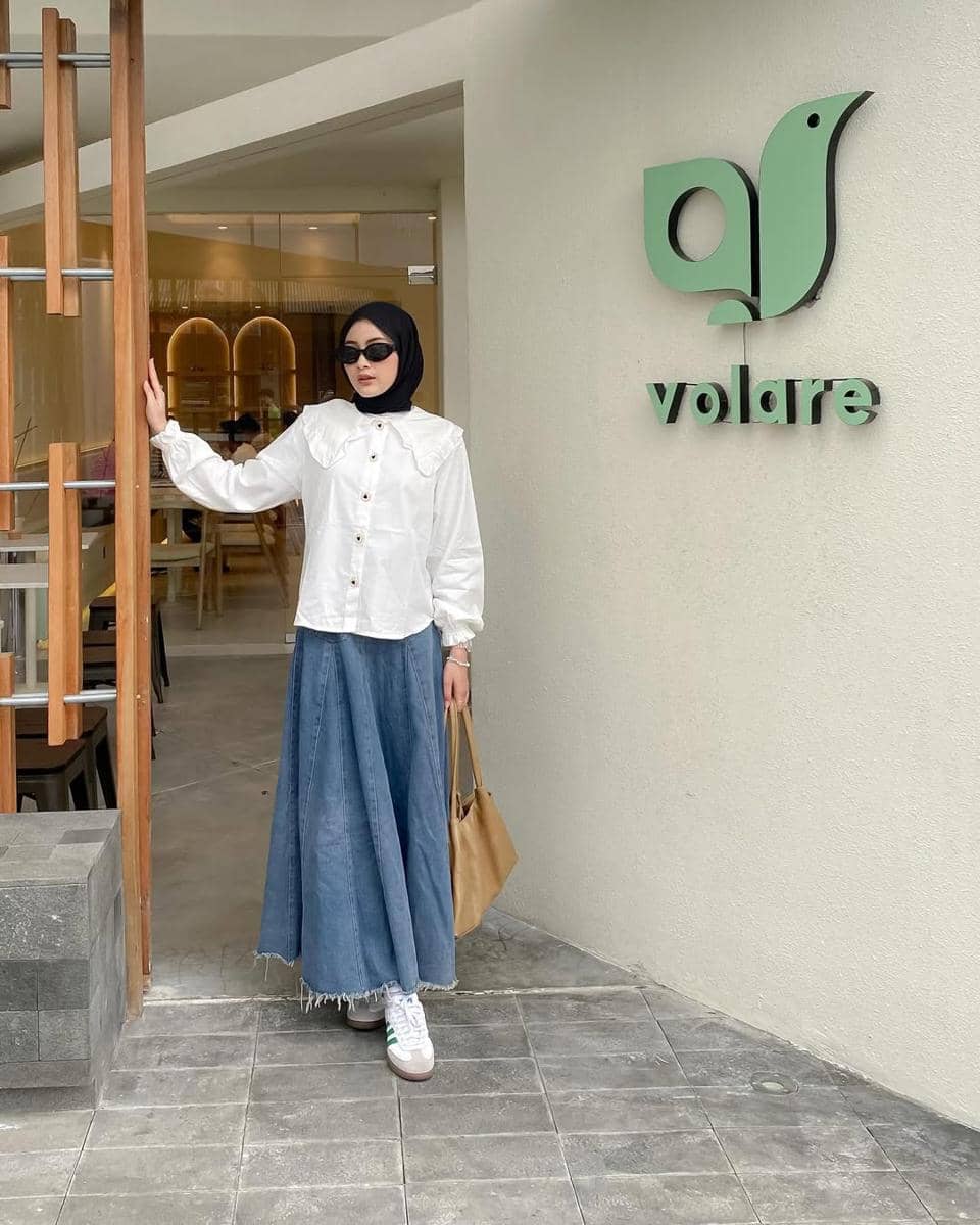 Seorang wanita berhijab mengenakan kemeja putih dan rok denim panjang dengan tas cokelat berdiri di depan kafe Volare.