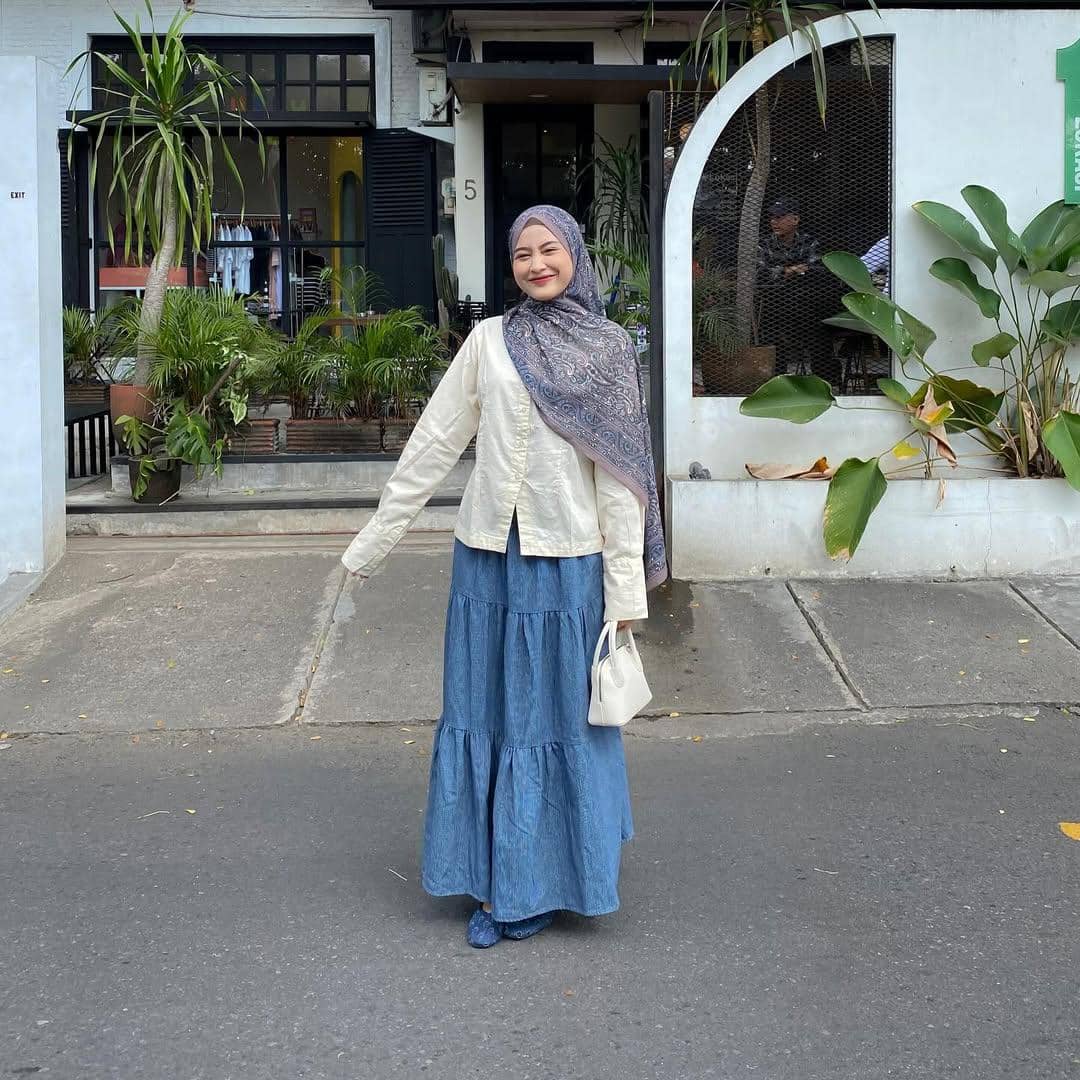 Seorang wanita berhijab bergaya kasual dengan rok denim panjang, atasan krem, dan tas kecil putih berpose di depan kafe modern.