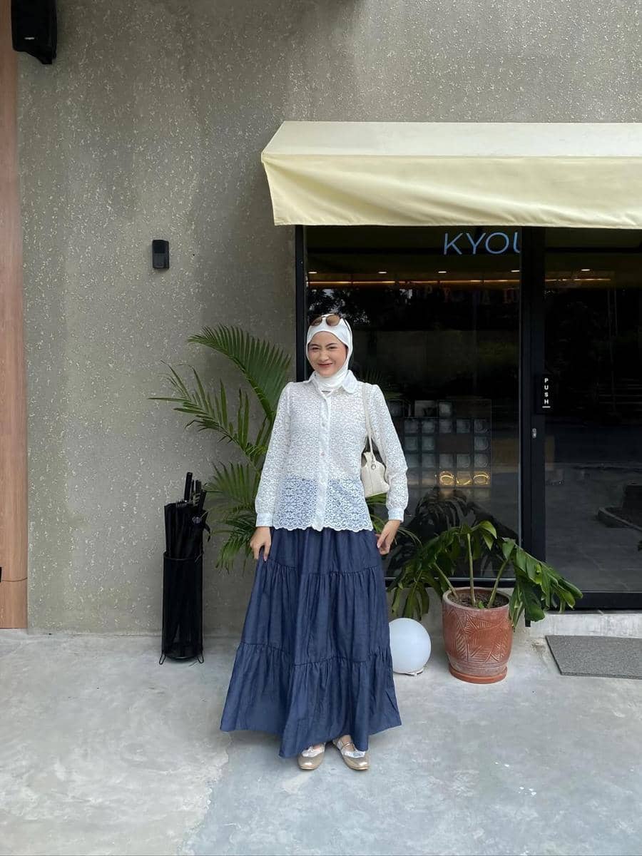 Seorang wanita berhijab mengenakan blus renda putih dan rok panjang biru tua berpose di depan kafe dengan tanaman hias di sekitarnya.
