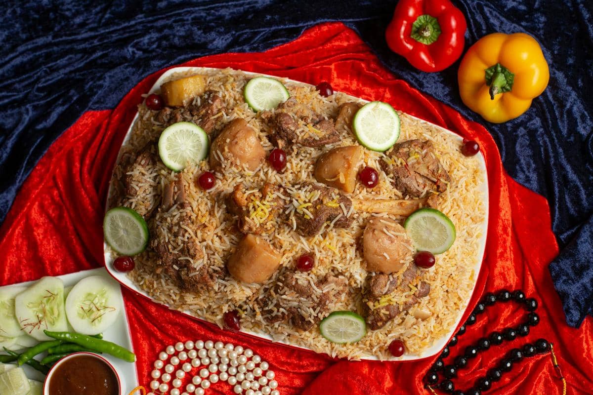Hidangan ayam biryani disajikan di atas piring besar dengan irisan jeruk nipis, nasi berbumbu, dan potongan ayam di atas kain merah dan biru.