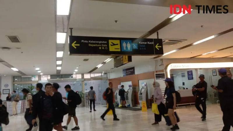 Penumpang berjalan di area dalam Bandara Sultan Hasanuddin dengan papan petunjuk arah menuju gerbang keberangkatan dan toilet.