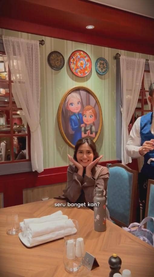 Seorang perempuan tersenyum sambil berpose di meja makan kapal pesiar bertema Disney dengan hiasan dinding karakter animasi Frozen di belakangnya.
