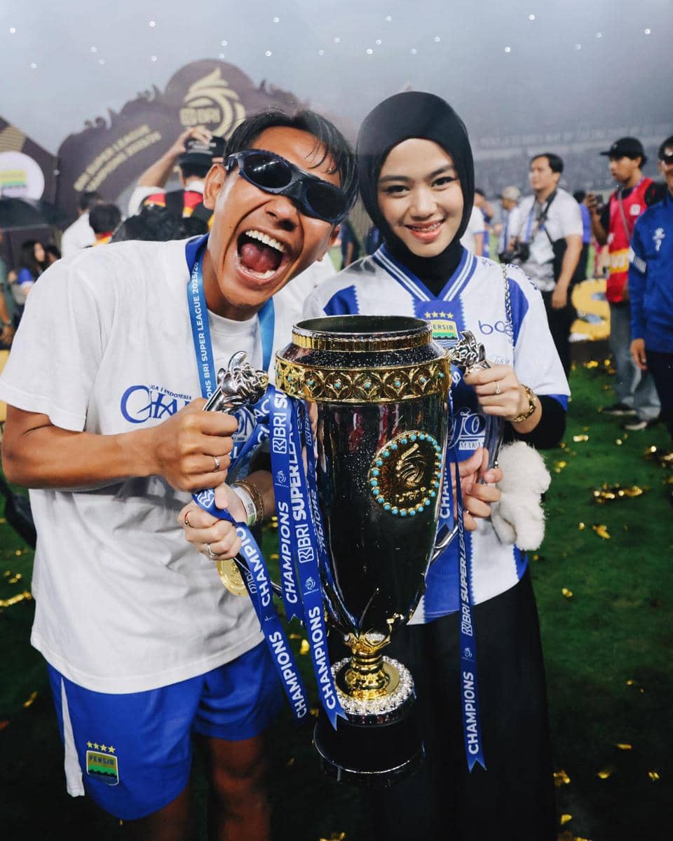Dua pendukung Persib Bandung berpose sambil memegang trofi juara di lapangan setelah kemenangan tim dalam kompetisi sepak bola.