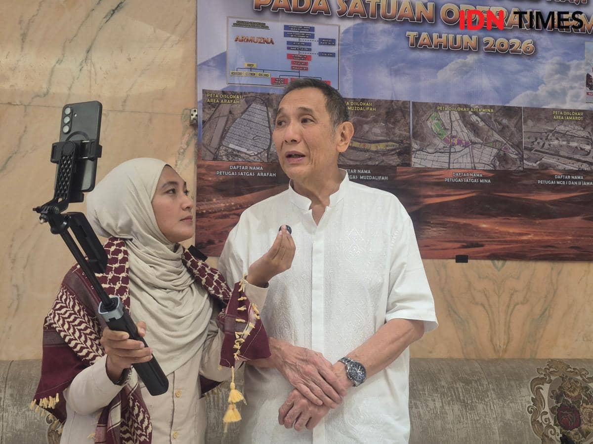 Seorang pria berbaju putih diwawancarai oleh perempuan berhijab yang memegang ponsel dengan tongkat selfie di depan poster bertema haji tahun 2026.