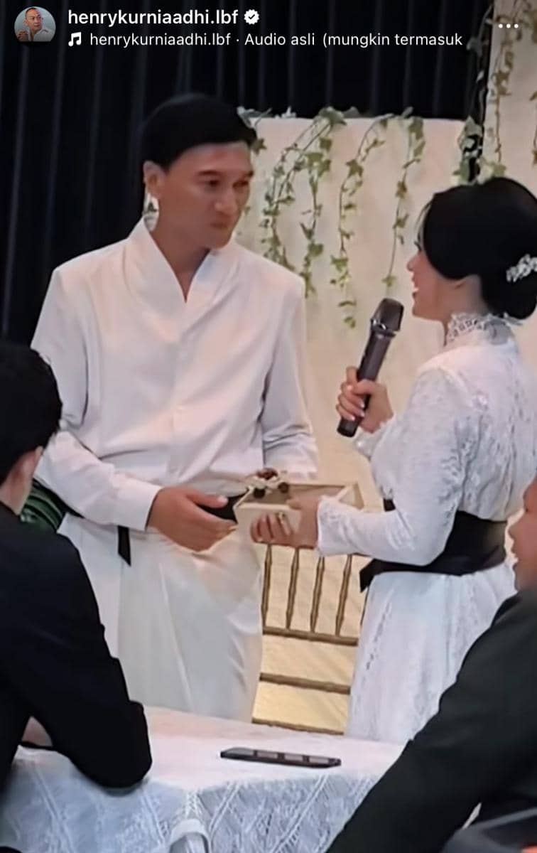 Seorang pria dan wanita mengenakan busana putih tradisional sedang bertukar cincin di acara pernikahan dengan dekorasi sederhana.