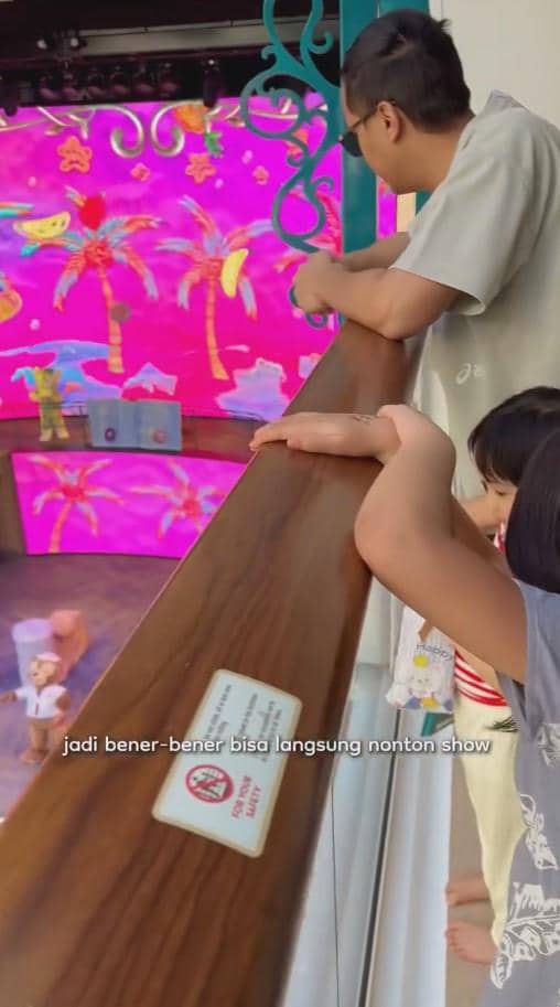 Seorang pria dan dua anak kecil menonton pertunjukan berwarna cerah bertema tropis di atas kapal pesiar Disney dari balkon.