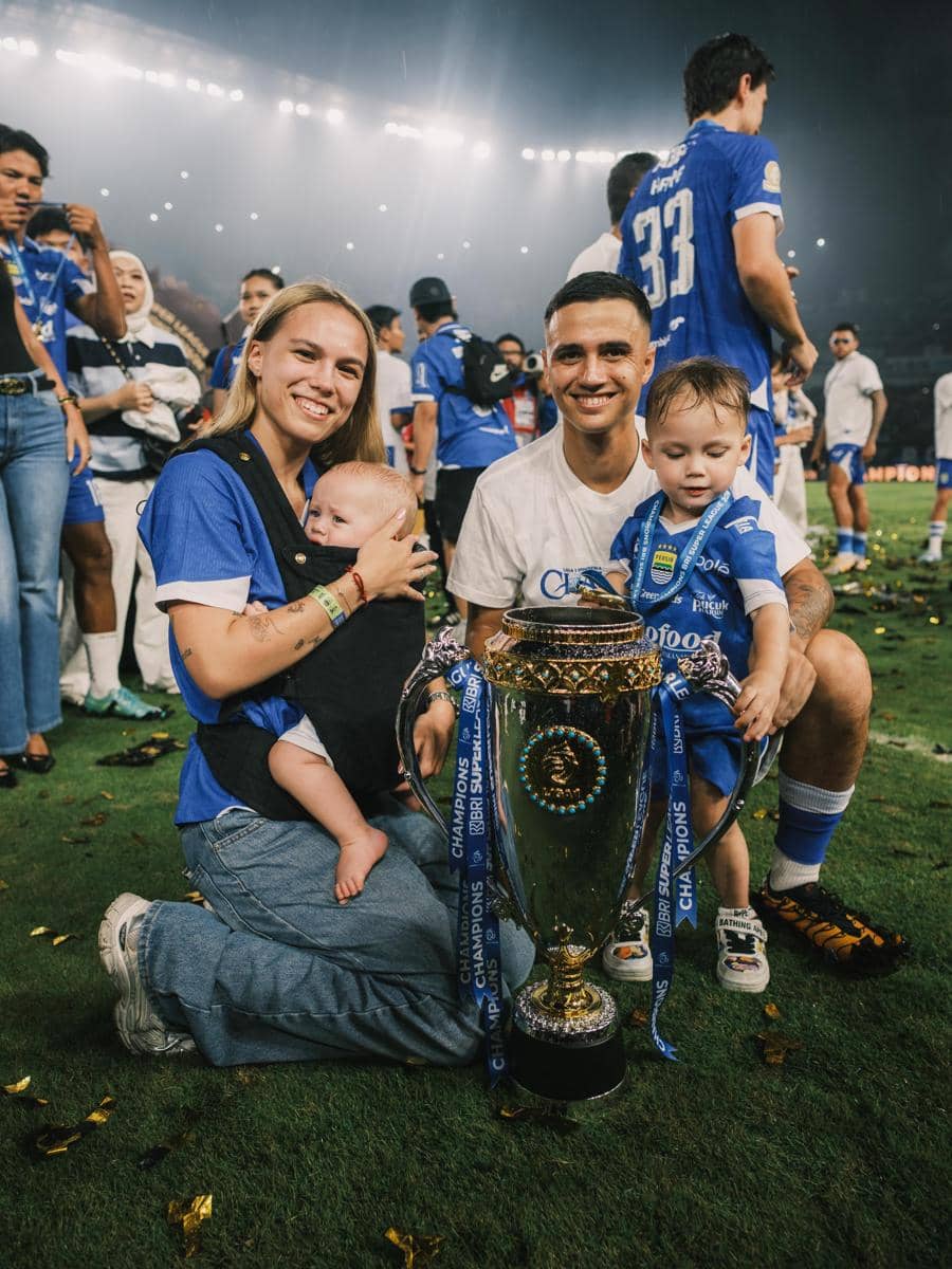Seorang pemain Persib Bandung berpose bersama keluarganya di lapangan sambil memegang trofi juara setelah pertandingan.