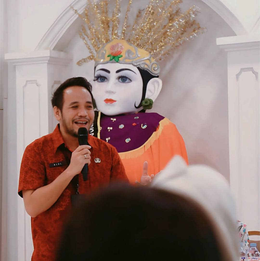 Seorang pria mengenakan kemeja merah berbicara menggunakan mikrofon di depan patung besar ondel-ondel dengan hiasan kepala emas.